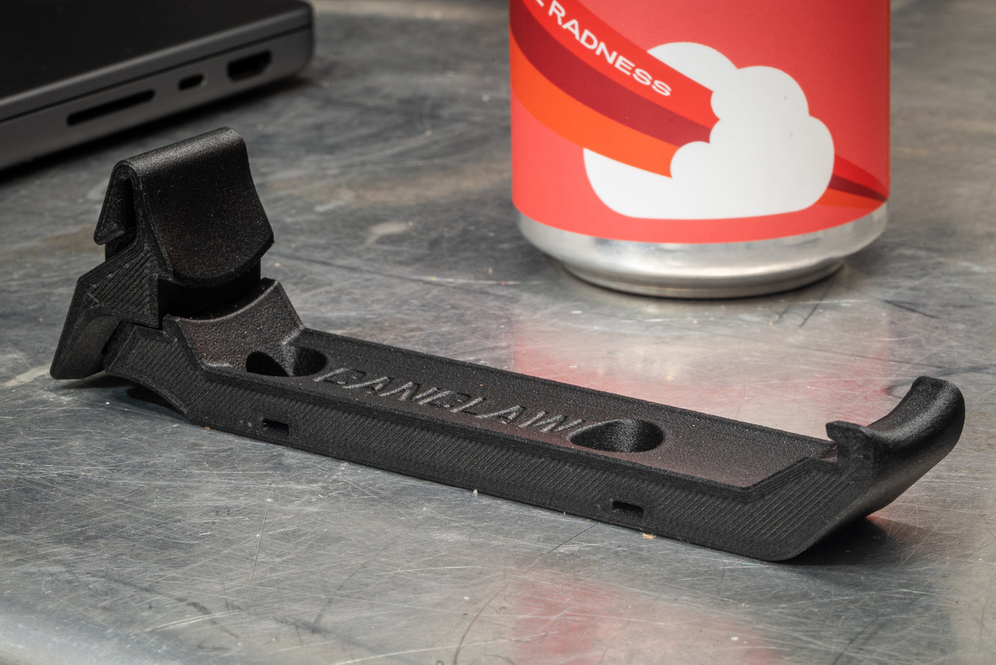 Image: The Canclaw, a lightweight and low-profile bike drink holder designed for standard 12oz (355ml) cans and 16oz (473ml) tall cans. It securely grips your beverage and can be attached using standard bottle cage bolt spacing or zipties. This product is 3D printed to order from ultra-strong carbon fiber nylon material and proudly made in Canada. Indoor Closeup.